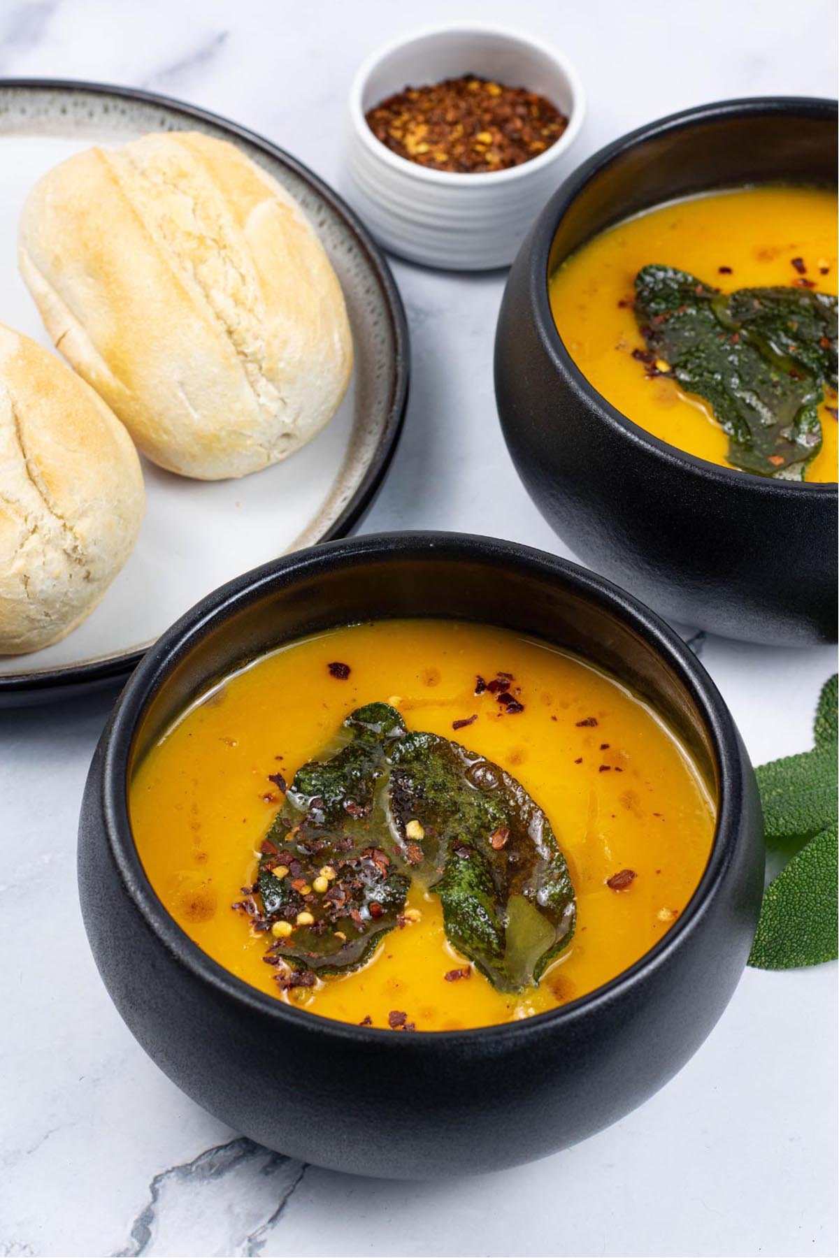 Two portions of squash and carrot soup with crispy sage leaves in black bowls with a plate of crusty rolls on the side.