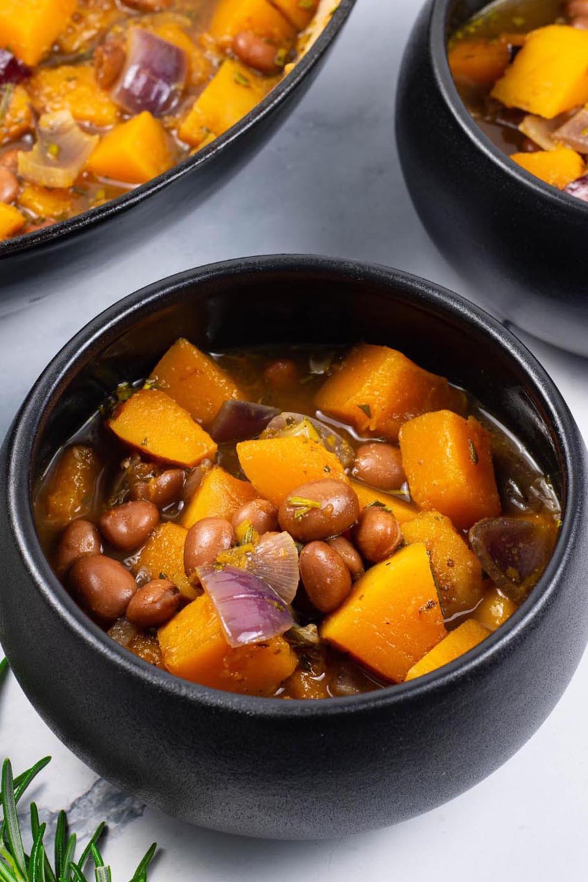 Two portions of sumptuous squash soup in black bowls with a sprig of rosemary on the side.