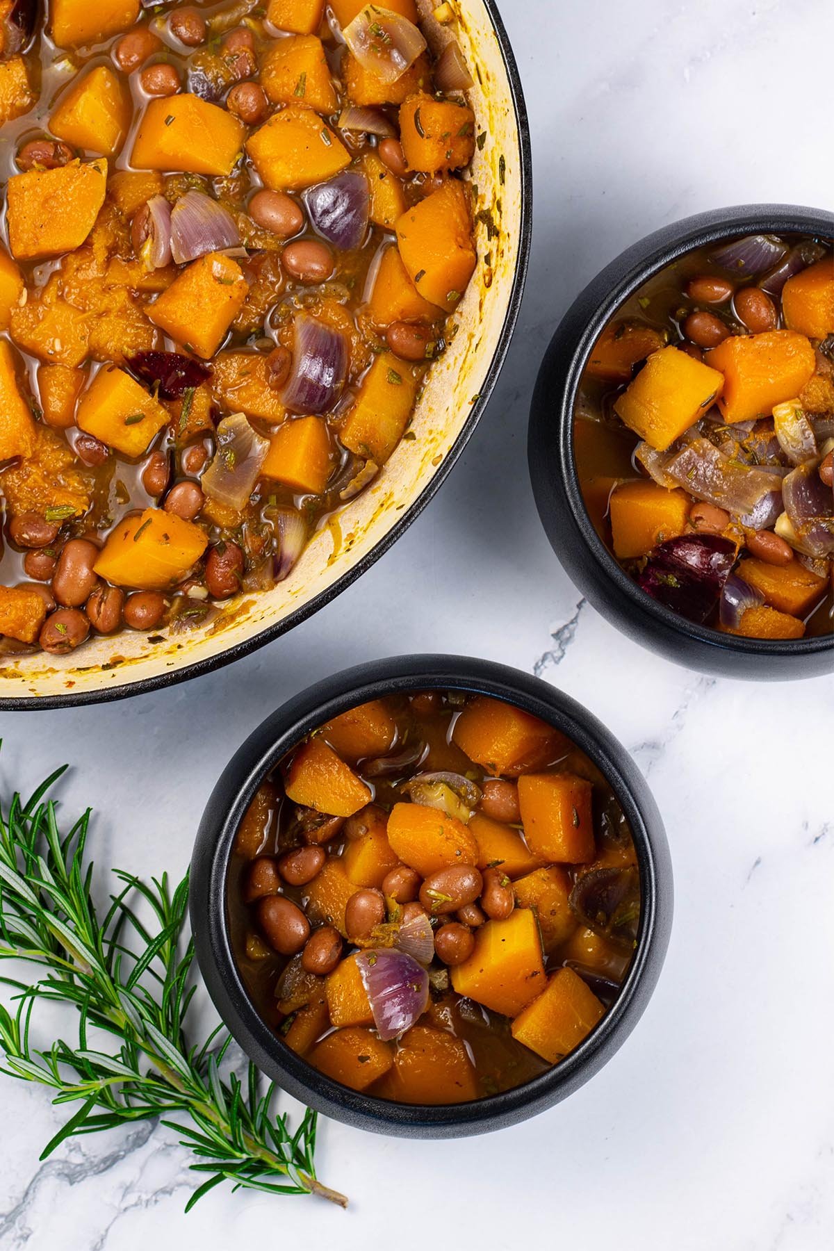 Two portions of sumptuous squash soup in black bowls with a sprig of rosemary on the side.