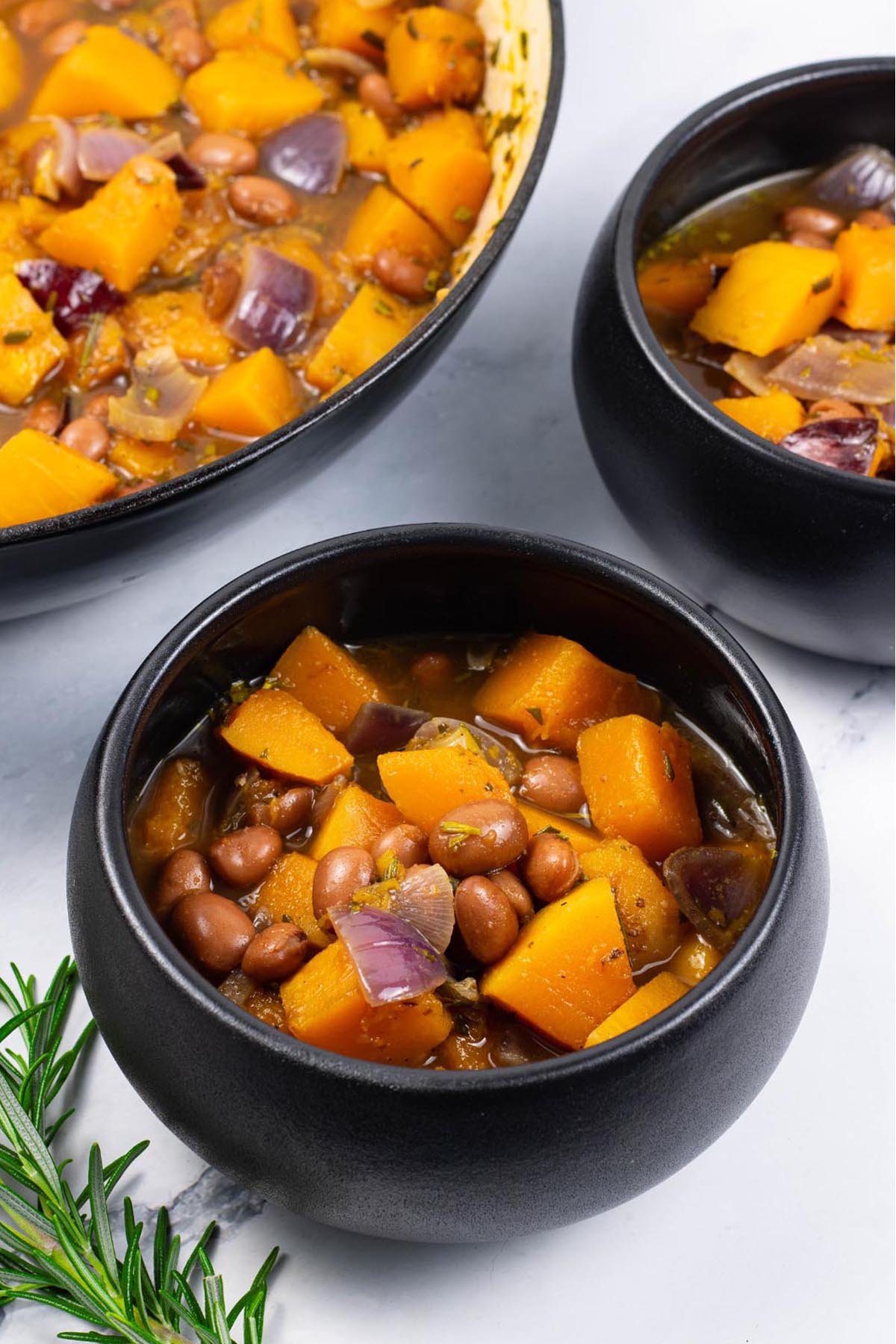 Two portions of sumptuous squash soup in black bowls with a sprig of rosemary on the side.