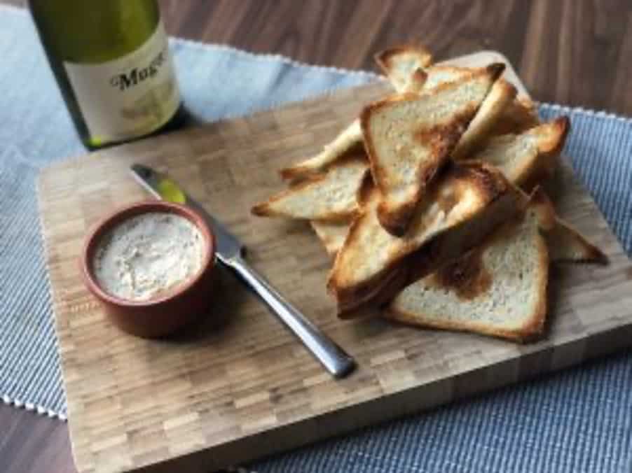 Pate in terracota ramekin with melba toast and knife on a wooden board and bottle of white wine on the side