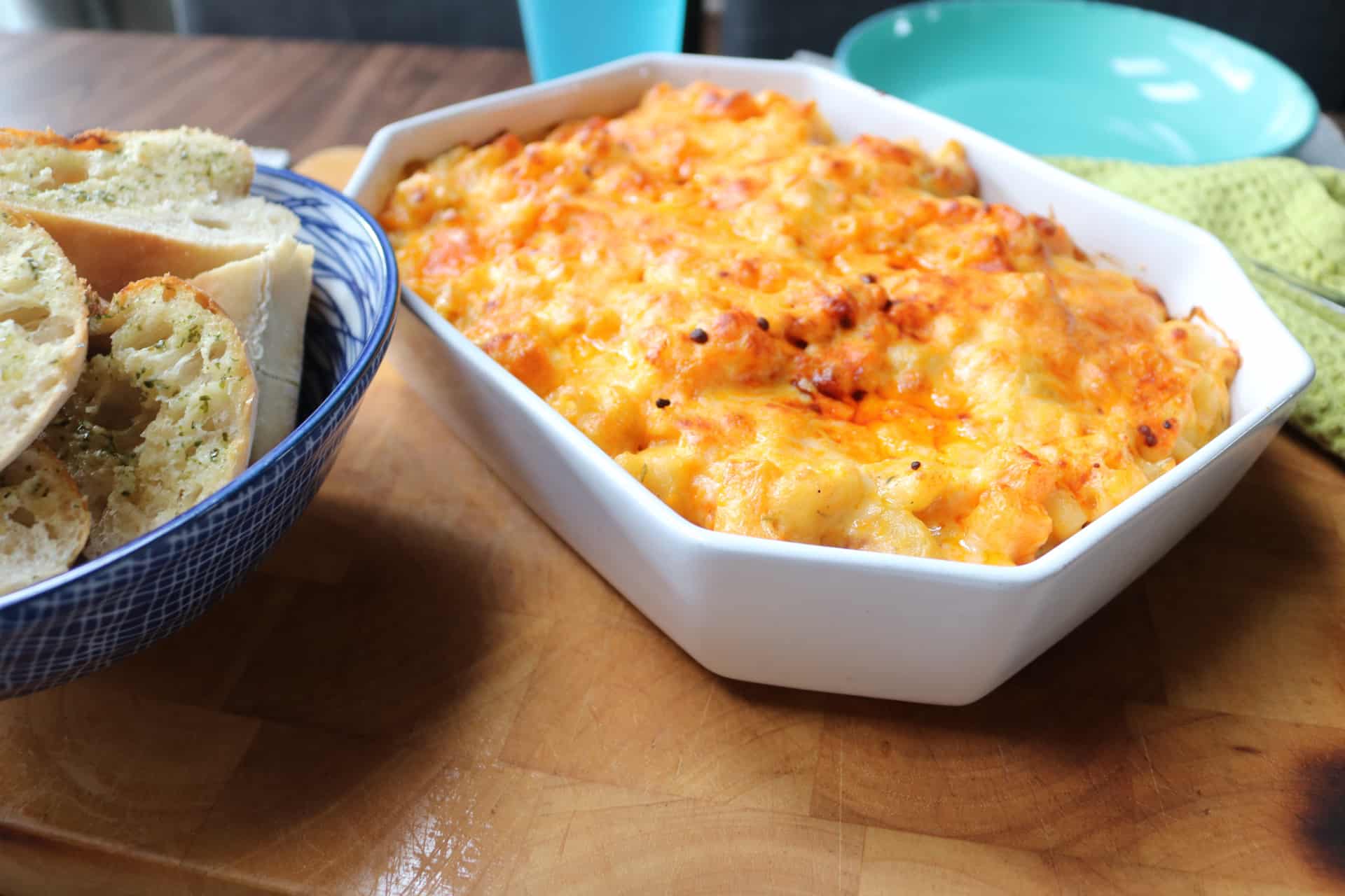 Butternut and bacon macaroni cheese in serving dish on large board with garlic bread in a bowl on the side, serving spoon and towel