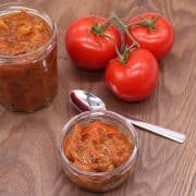Spicy tomato relish in jar and small bowl with tomatoes and spoon on wooden board