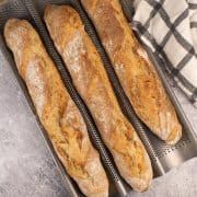 Three baguettes in a baguette baking tray beside black and white towel