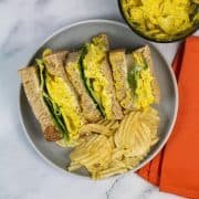 Coronation chicken sandwiches on a grey plate with crinkle crisps. Small bowl of extra sandwich filler and orange napkin in background