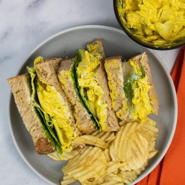 Coronation chicken sandwiches on a grey plate with crinkle crisps. Small bowl of extra sandwich filler and orange napkin in background