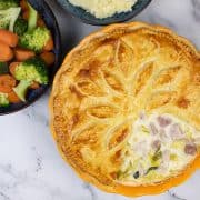 Creamy chicken, gammon and leek pie in a round pie dish with bowls of veg and mash in the background