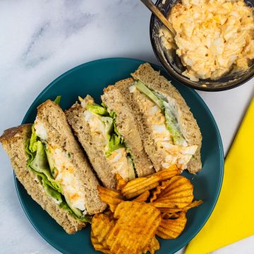 Egg mayonnaise sandwiches on a dark green plate with crinkle crisps. Small bowl of extra sandwich filler and yellow napkin in background