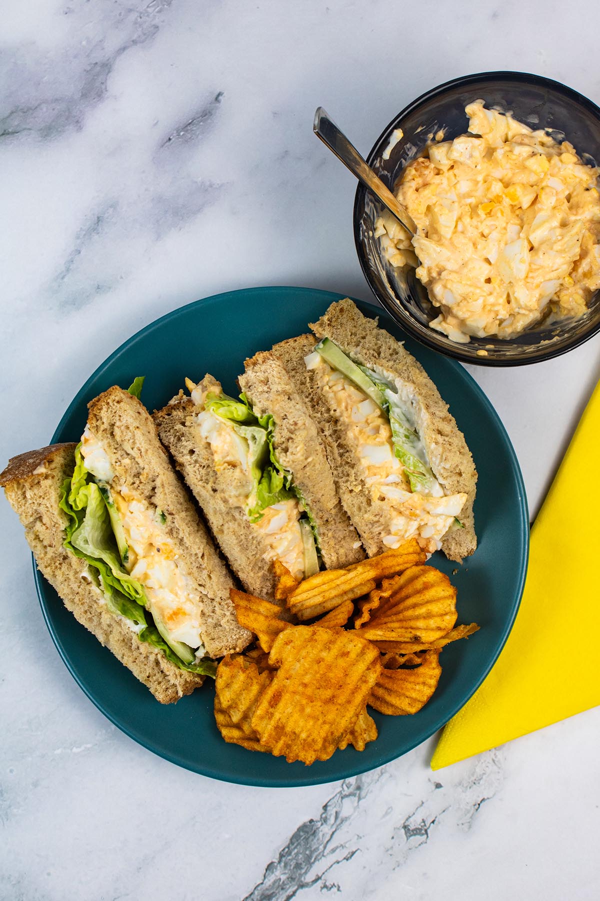 Egg mayonnaise sandwiches on a dark green plate with crinkle crisps. Small bowl of extra sandwich filler and yellow napkin in background