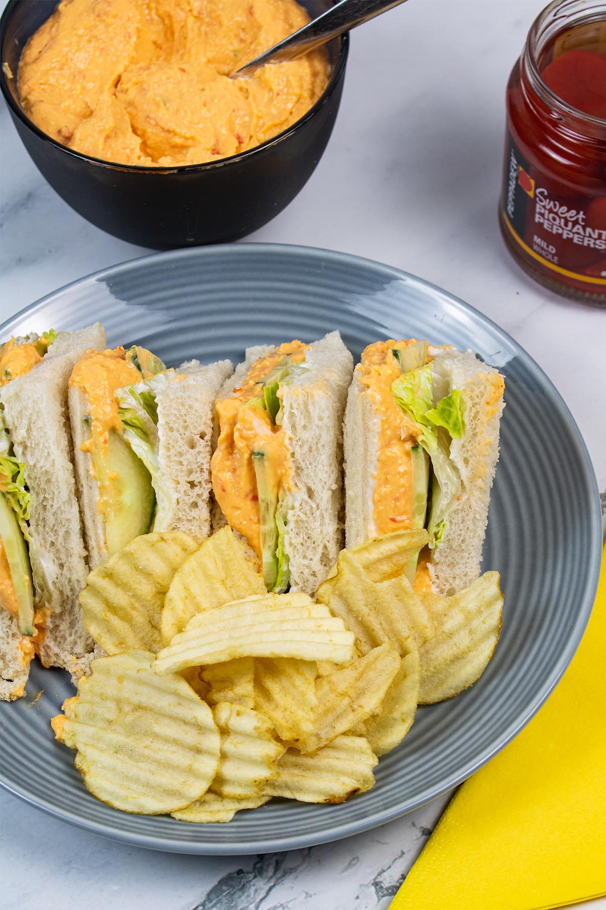 Pimento cheese sandwiches on a grey plate with crinkle crisps. Small bowl of extra sandwich filler, jar of piquante peppers and yellow napkin in background