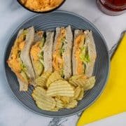 Pimento cheese sandwiches on a grey plate with crinkle crisps. Small bowl of extra sandwich filler, jar of piquante peppers and yellow napkin in background