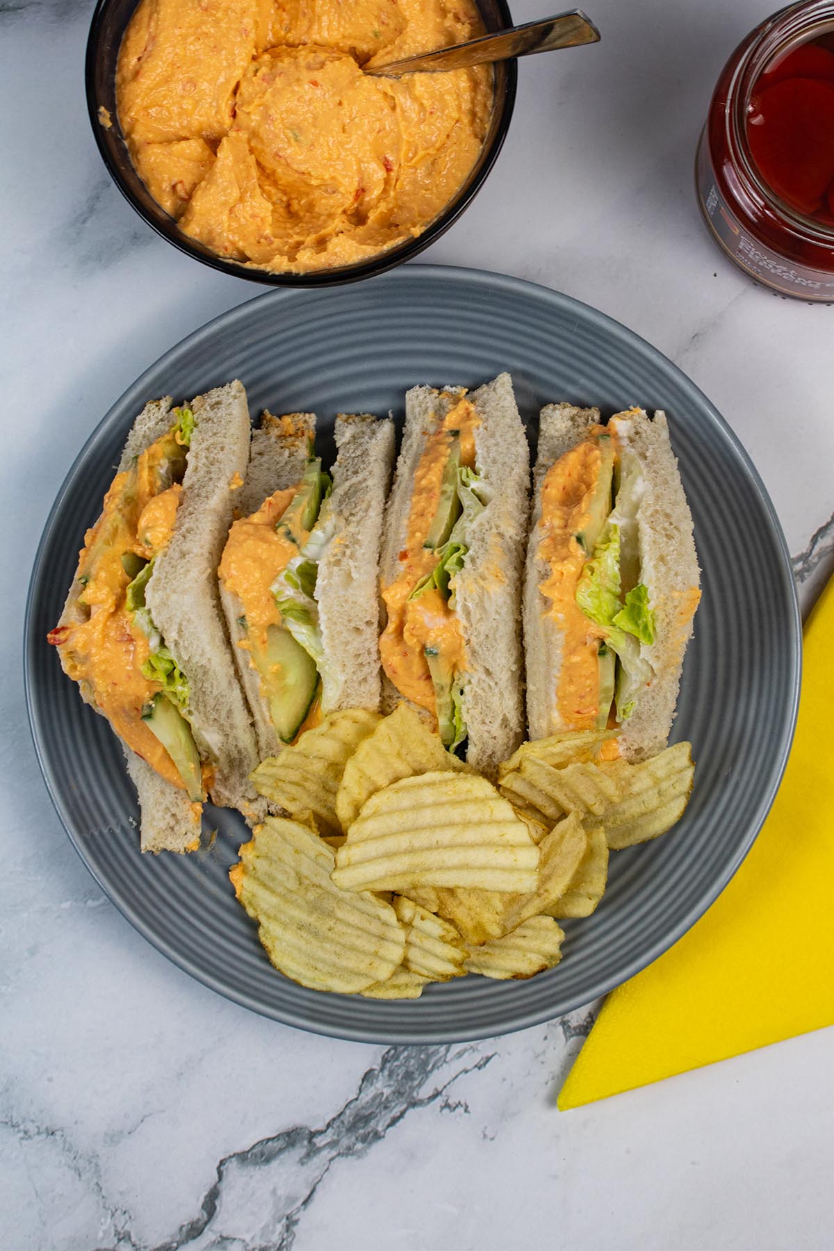 Pimento cheese sandwiches on a grey plate with crinkle crisps. Small bowl of extra sandwich filler, jar of piquante peppers and yellow napkin in background