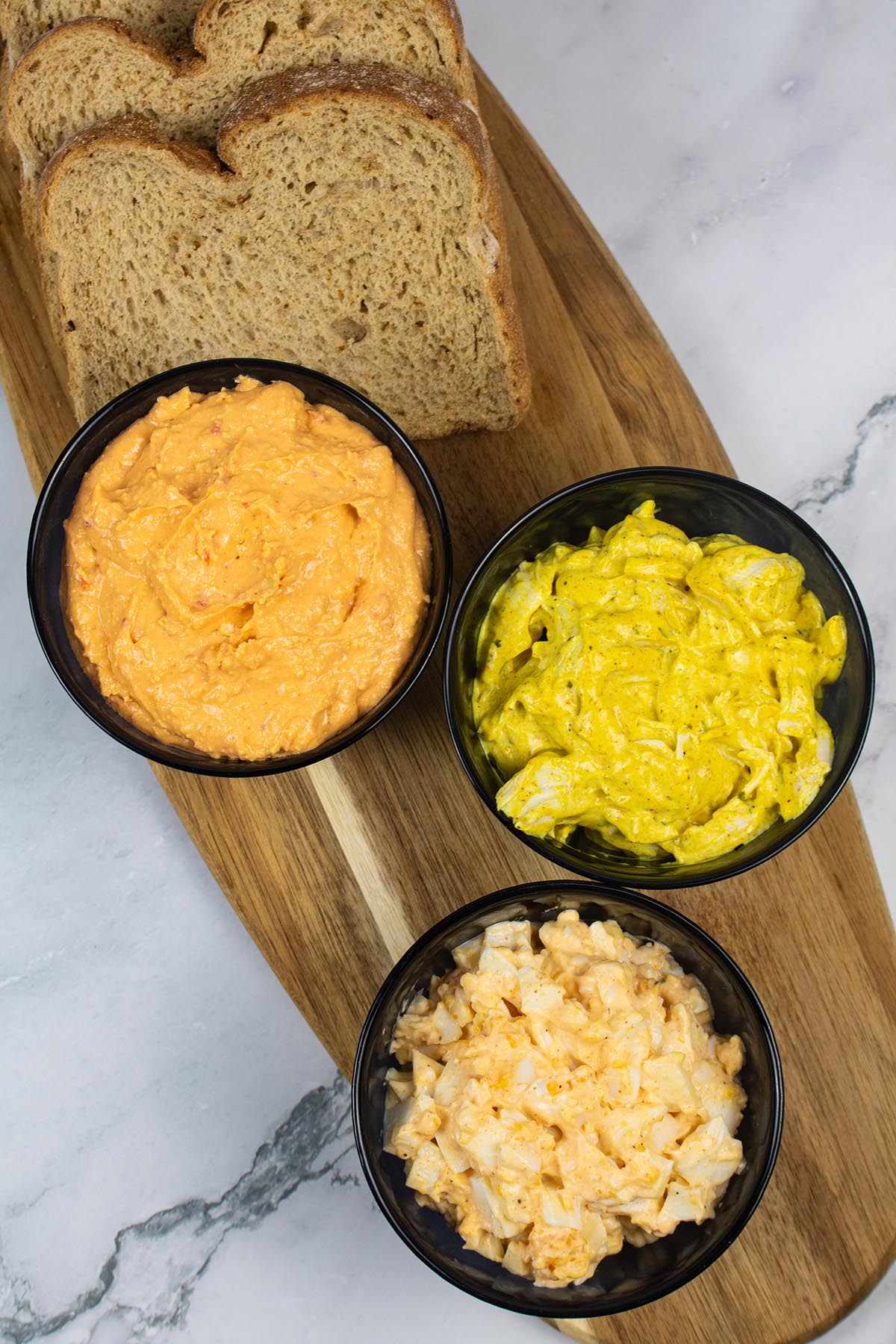 Small black bowls of egg mayo, pimento cheese and coronation chicken sandwich fillers on a board with sliced brown bread