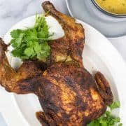 Tandoori roast chicken on a white platter with coriander garnish and sauce in a grey jug on the side.