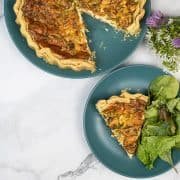 Quiche on dark green plates - one with a slice and salad, with a bunch of wild garlic and chive flowers at the side.