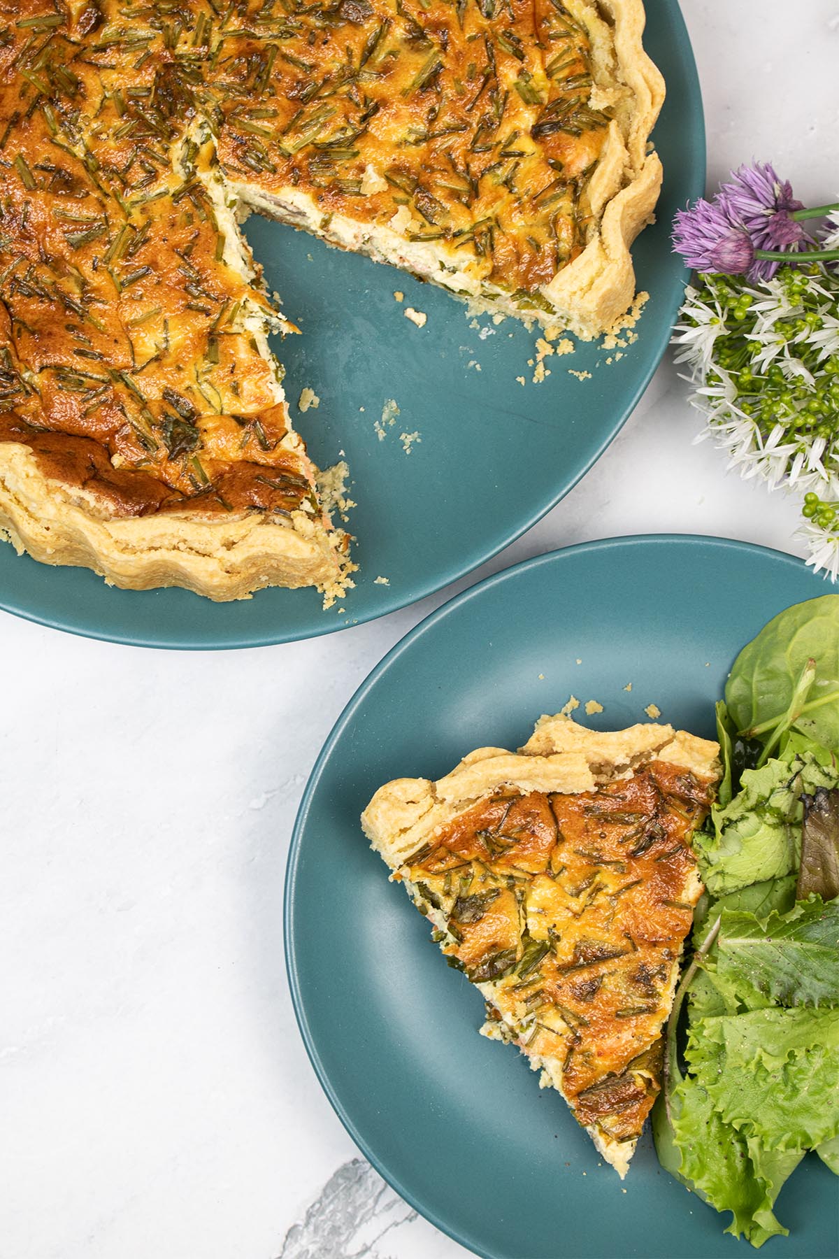 Quiche on dark green plates - one with a slice and salad, with a bunch of wild garlic and chive flowers at the side.