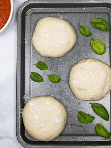 3 balls of pizza dough on baking tray scattered with basil leaves and dish of pizza sauce in the background