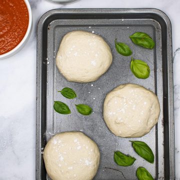 3 balls of pizza dough on baking tray scattered with basil leaves and dishes of pizza sauce and grated mozzarella in the background