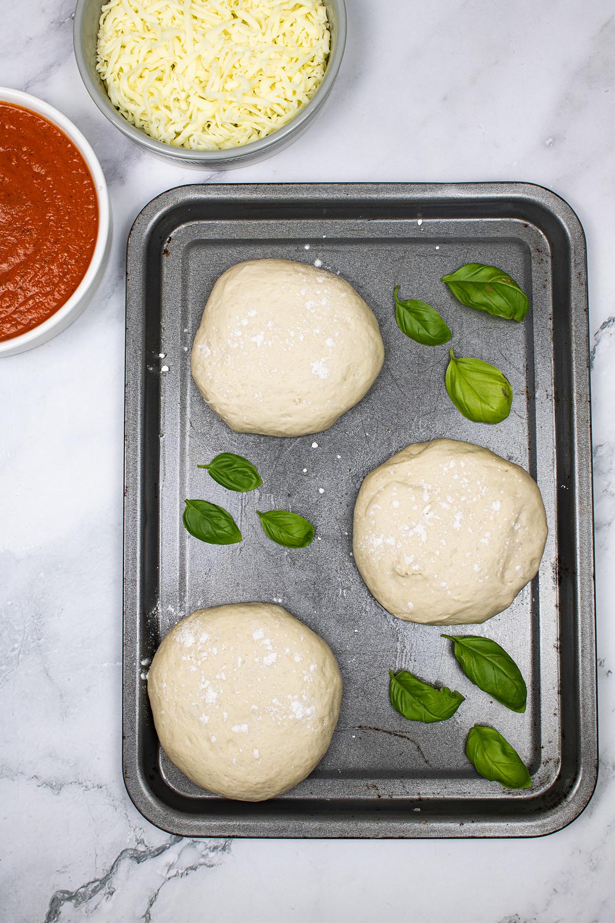 3 balls of pizza dough on baking tray scattered with basil leaves and dishes of pizza sauce and grated mozzarella in the background
