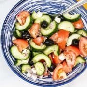 Greek salad in a blue and white striped bowl with serving spoon and yellow napkin in background