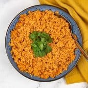 Red rice in a blue patterned serving bowl with spoon, garnished with coriander and with a mustard yellow napkin on the side