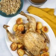 Roast chicken on a white platter with roast potatoes, a teal bowl of stuffing and grey jug of gravy in the background.