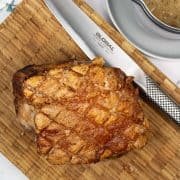 Cooked roast pork with crispy crackling on a wooden board with Global carving knife. Cider and onion gravy in a jug on the side.