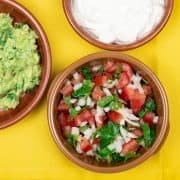 Pico de gallo salsa in a terracotta dish on top of a yellow napkin. Dishes of guacamole and sour cream in the background.