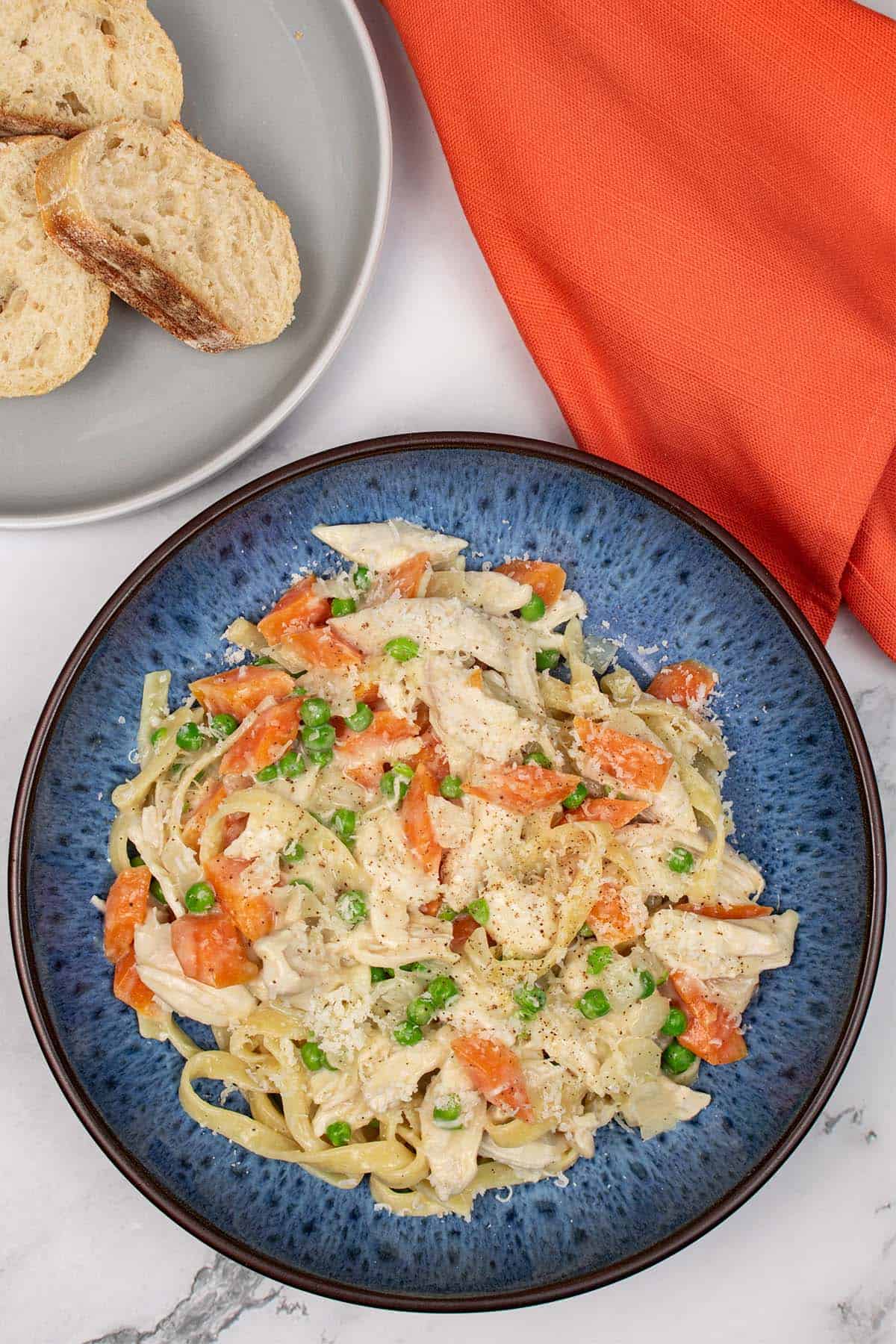 Creamy chicken and vegetable tagliatelle in a blue pattered bowl with slices of crusty bread on a grey side plate, and orange napkin on the side.