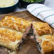 Salmon en croute on a board cut open with a teal and white striped towel and bowls of mashed potatoes and green veg on the side.