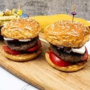 2 lamb burgers in seeded brioche buns on a wooden board with a bowl of feta and oregano fries on the side.