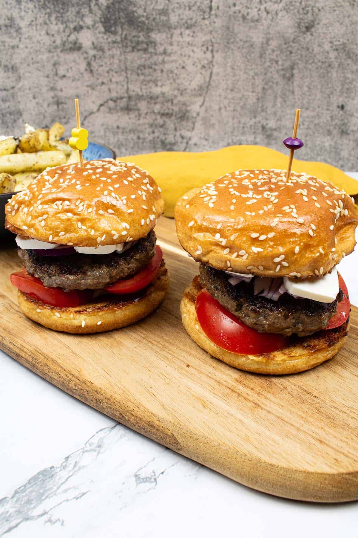 2 lamb burgers in seeded brioche buns on a wooden board with a bowl of feta and oregano fries on the side.