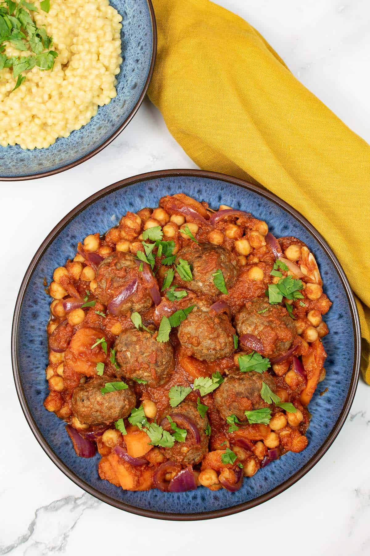 Lamb meatball tagine in a blue patterned bowl, with a bowl of couscous and mustard napkin on the side.