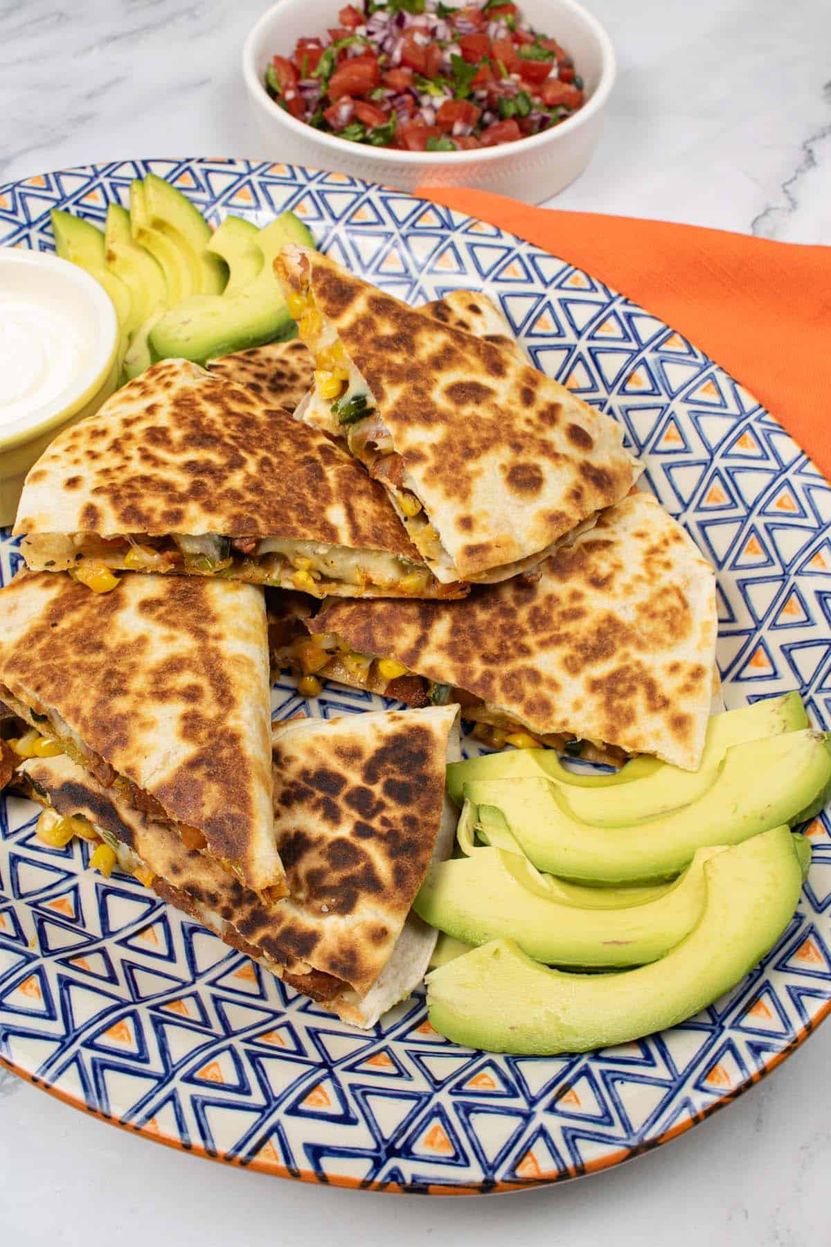 Quesadillas with sweet potatoes, chorizo and sweetcorn on a platter with slices of avocado, a ramekin of sour cream, and a dish of Pico De Gallo salsa in the background.