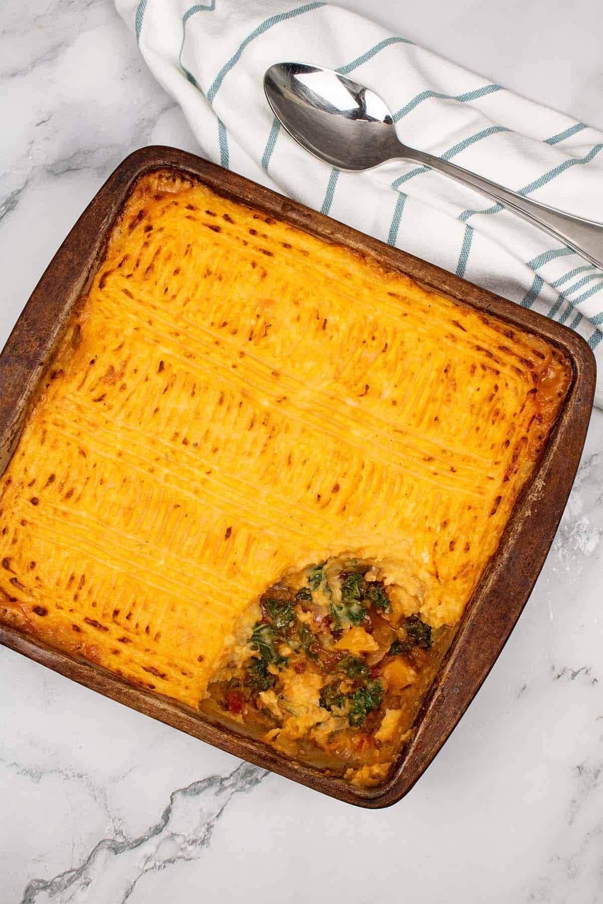 Vegetable and lentil cottage pie in a square stoneware dish with a serving spoon and teal and white towel on the side.