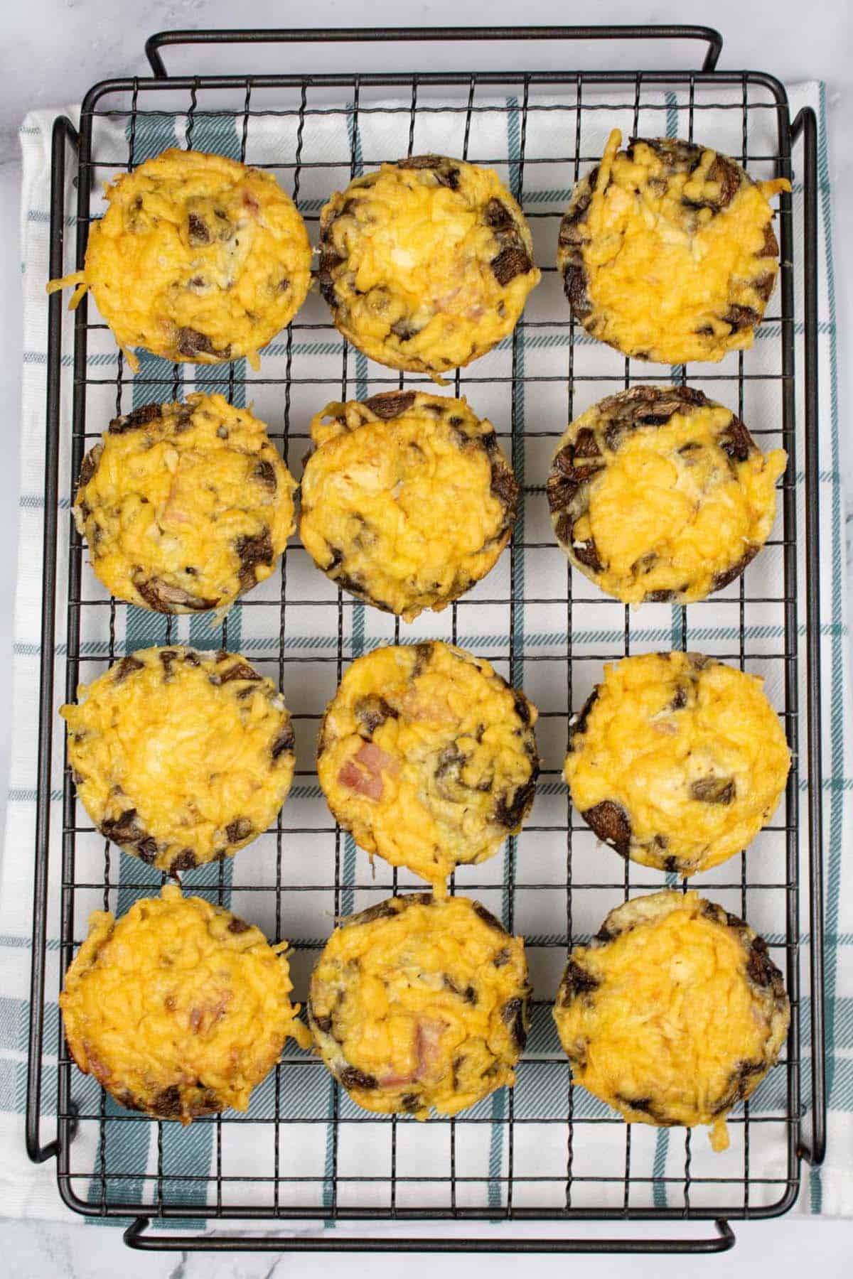 12 cheese, ham and mushroom omelette muffins on a cooling rack, on top of a teal and white checked towel.