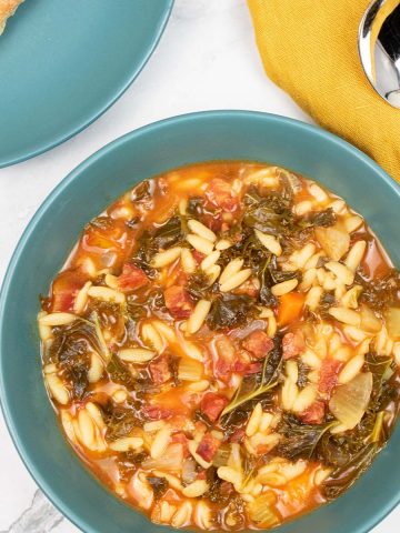Chorizo and orzo soup in a teal bowl with a side plate and ciabatta roll, soup spoon and mustard napkin on the side.