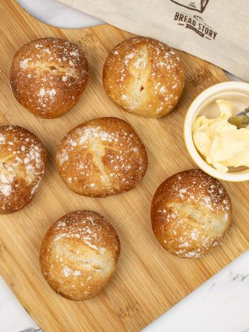 Crusty white dinner rolls on a board with a dish of butter with butter knife. Bread bag in the background.