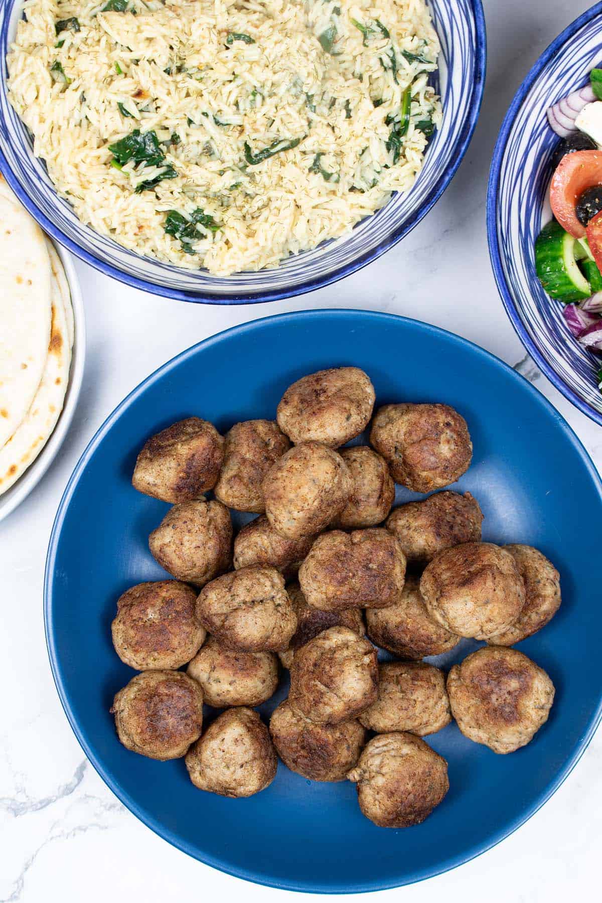 Greek lamb meatballs made in the Thermomix in a blue bowl with Greek rice with spinach, Greek salad and flatbreads on the side.