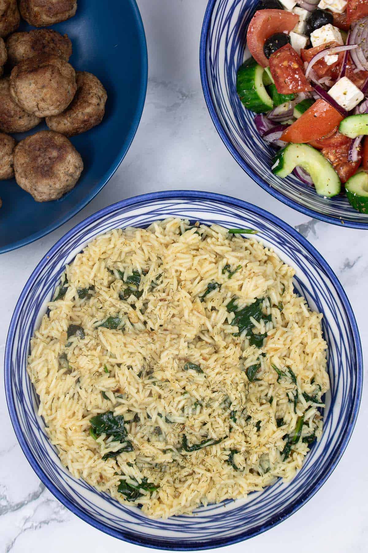 Greek rice with spinach in a blue and white patterned bowl, with bowls of lamb meatballs and Greek salad in the background.
