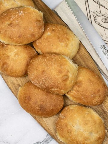 Homemade ciabatta bread rolls on a board with a bread knife and bread bag on the side.