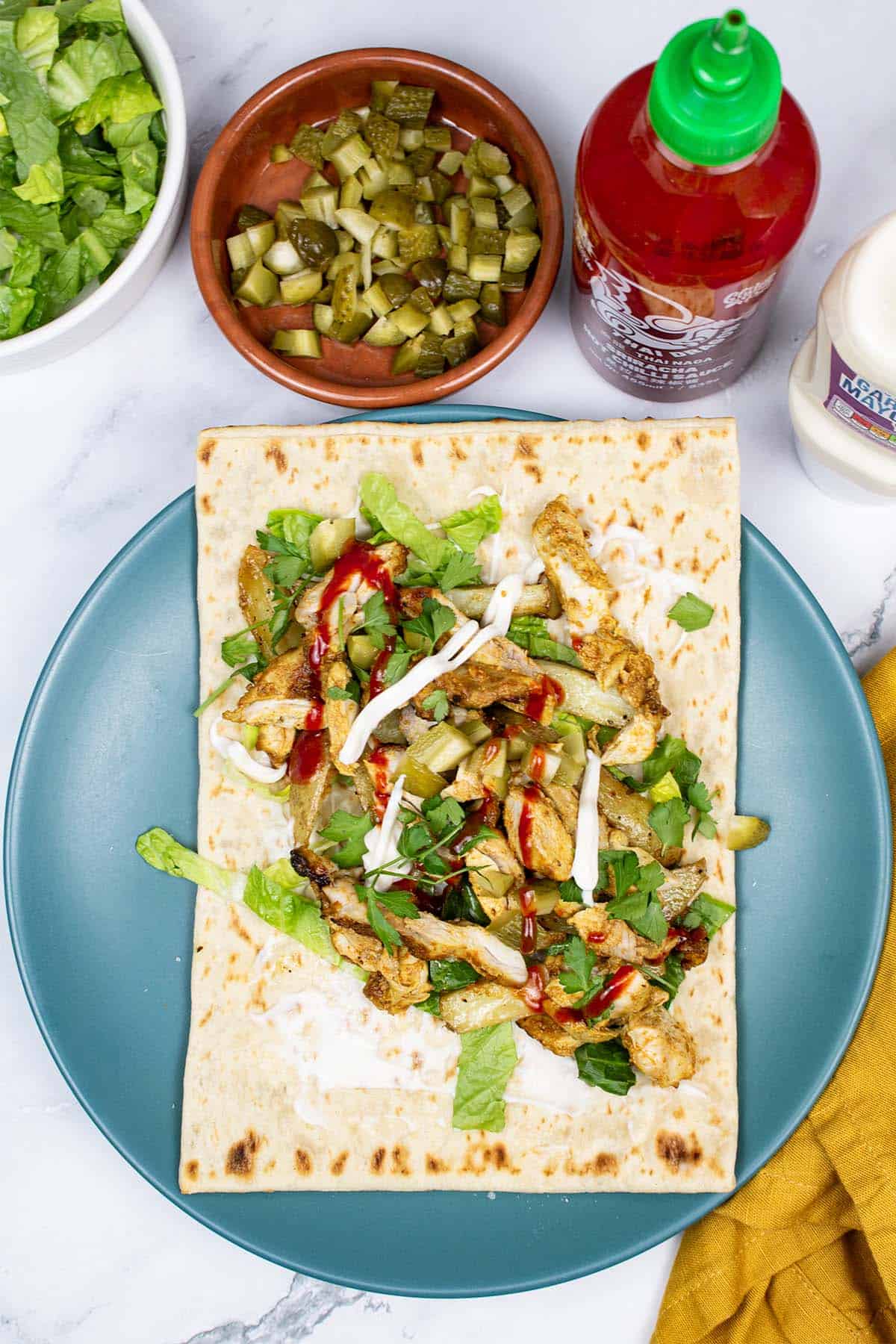 Kebab king shawarma assembled on a teal dinner plate with dishes of shredded lettuce, chopped gherkins and bottles of sriracha and garlic mayo on the side.