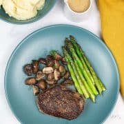 Perfect fillet steak on a teal dinner plate with mushrooms and asparagus. A bowl of horseradish mash and small jug of whisky sauce and a mustard napkin on the side.
