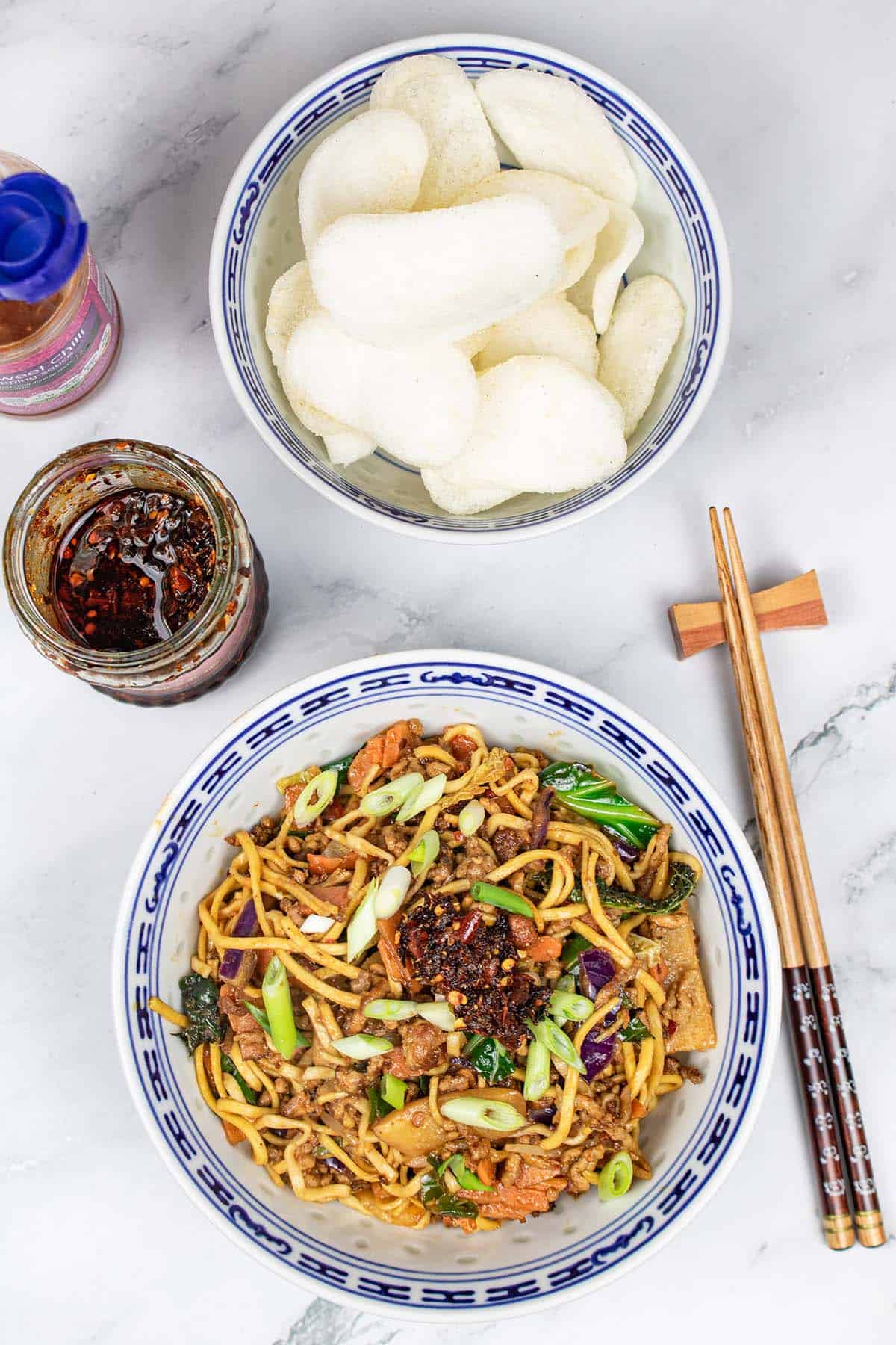 Pork noodle stir-fry in and oriental bowl with a bowl of prawn crackers, jar of crispy chilli in oil, bottle of sweet chilli sauce and chopsticks on the side.