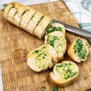 Quick and easy garlic baguette on a board with 5 slices cut, a bread knife and teal and white checked towel on the side.