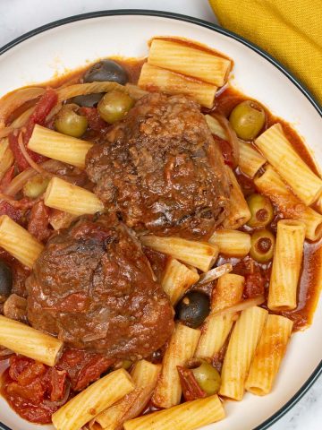 A portion of slow cooker chicken cacciatore served with rigatoni in a black rimmed pasta bowl with a mustard napkin on the side.