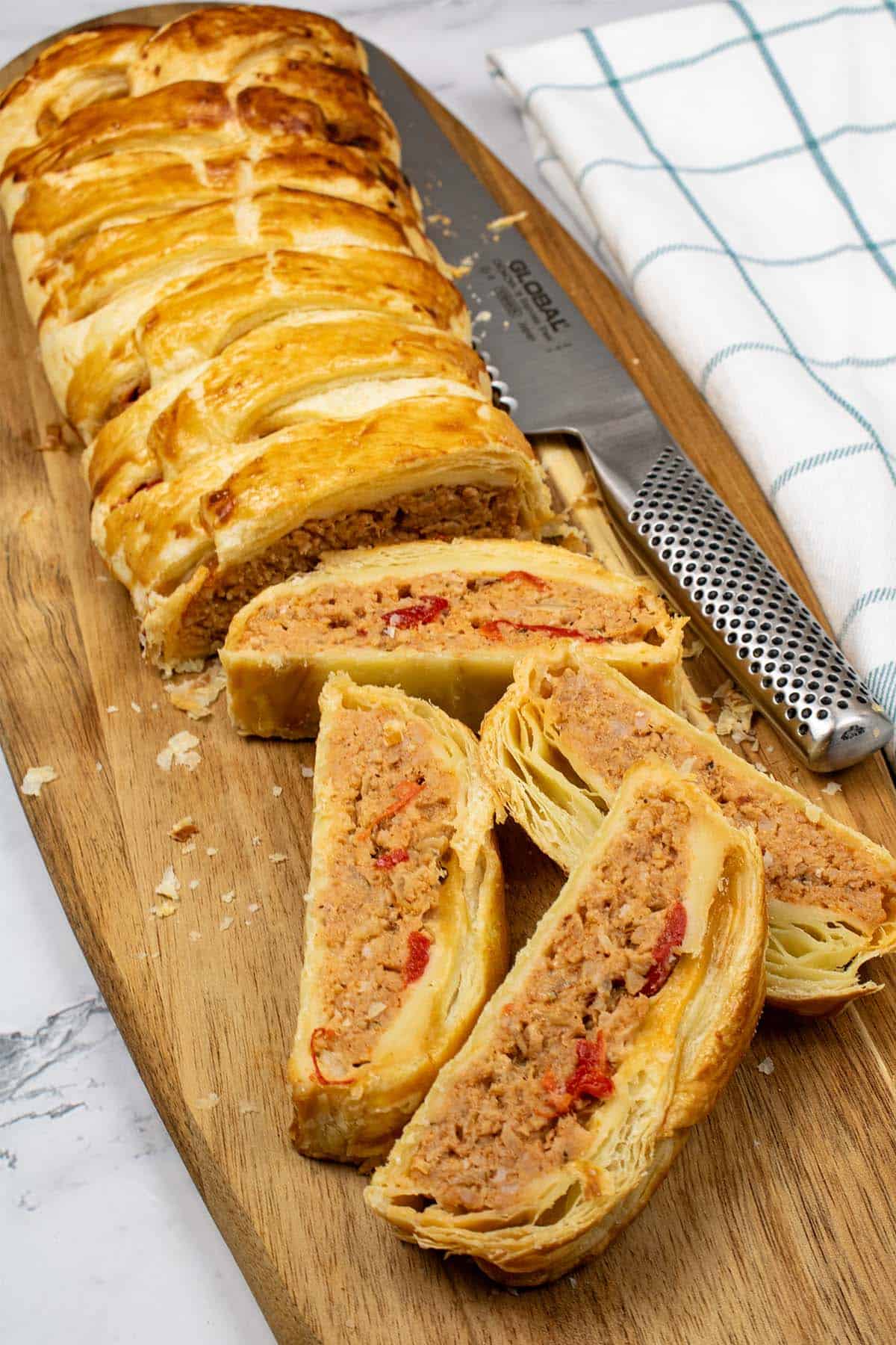 Spanish-flavoured sausagemeat plait made in Thermomix on a wooden board with 4 pieces sliced, a bread knife and teal and white checked towel on the side.