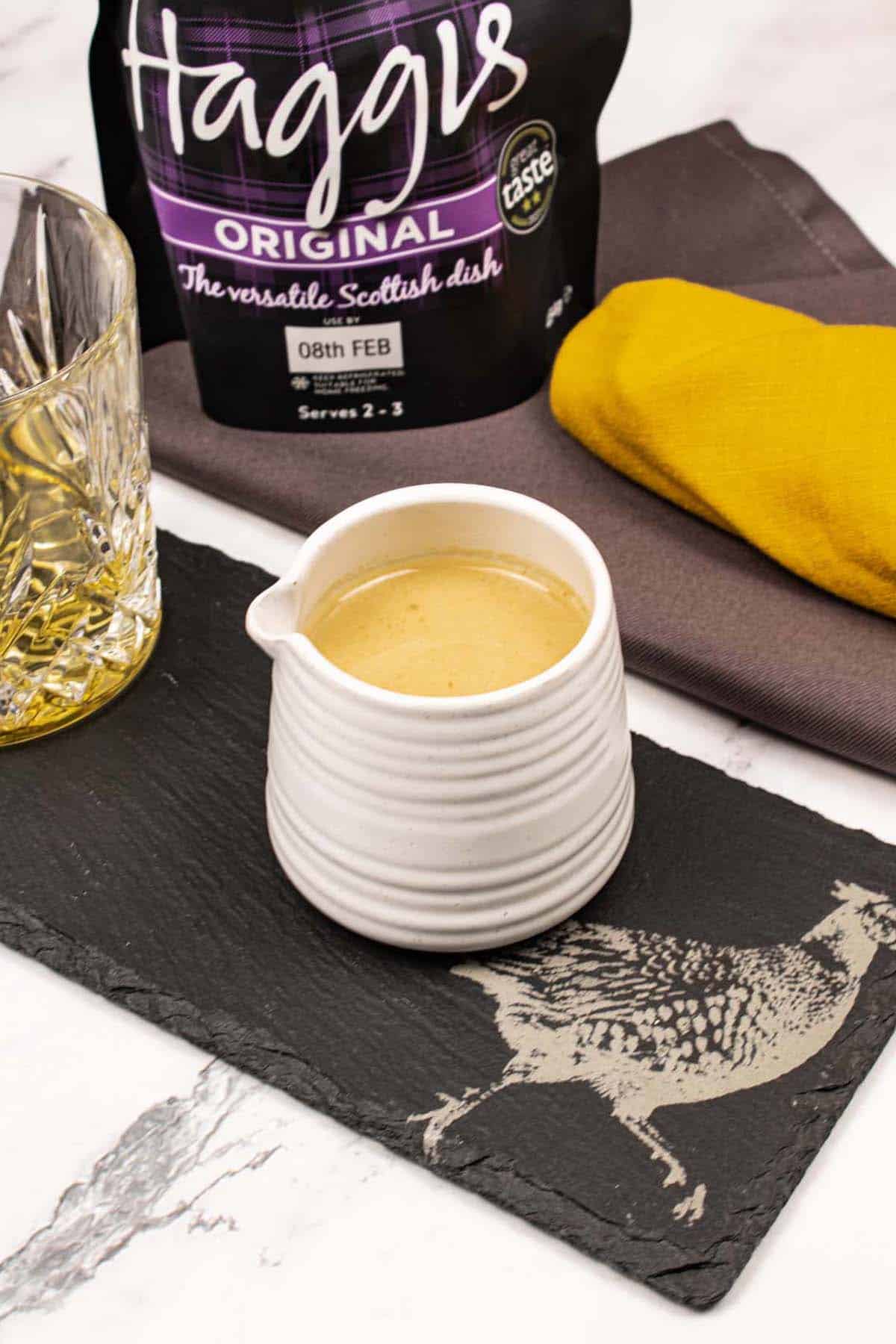 Whisky sauce made in Thermomix in a small cream ribbed jug sitting on a black slate with a dram of whisky. A packet of haggis and charcoal and mustard napkins in the background.