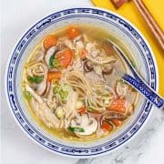 Chinese chicken noodle soup in a blue and white oriental bowl with spoon. A yellow napkin and chopsticks on the side.
