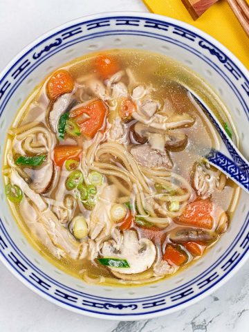 Chinese chicken noodle soup in a blue and white oriental bowl with spoon. A yellow napkin and chopsticks on the side.
